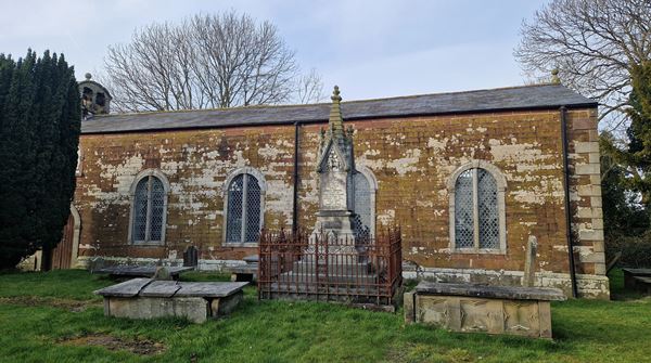 Photo of a small chapel built of yellowish stone. The view is from the side, showing 4 windows and a wooden door, all with a semicircular arched shape. There's a small bell at one end of the peak of the roof. In the foreground there are several tombstones and grave monuments, and a large yew tree..